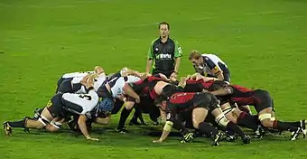 Two opposing formations of eight men, in white and black to the left, red and black to the right, push against each other in a crouched position; behind them stands another player and the referee