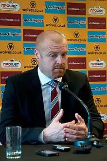 A football manager is sitting behind a microphone, while his hands are cupped