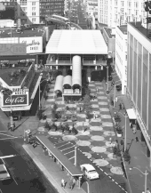 An overhead view of the monorail terminal with a large roof, and a stairway leading down to a checker-patterned plaza with a car lane and bus shelter