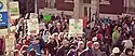 Marchers, most wearing jackets, walk along a street in front of a brick building, carrying signs.