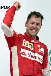 A man in his late twenties wearing red racing overalls with sponsors logos. He is holding his right arm in the air to celebrate winning a motor race and is smiling