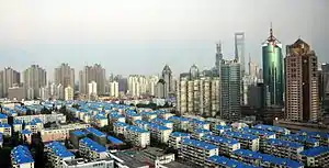 A cityscape seen from a height. In the foreground are low-rise buildings with blue roofs, giving way at the rear to higher-rising towers of different shapes and materials.