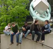 five young people sitting on stone wall in front of modern sculpture and tree