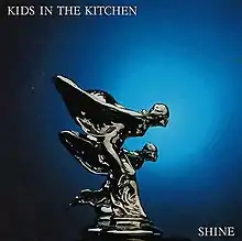 The album's name is seen at the top left, styled in white lettering and all caps over dark blue background. Two winged, female figures are in the middle of the field with a chrome finish. They are striving towards the right as if racing each other. They are mounted on a plinth similar to a car's hood ornament. At the bottom right is the album's name, again in all caps, white lettering.