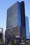 Ground-level view of a blue and black, rectangular, glass high-rise; one facade is covered in slightly protruding vertical stripes.