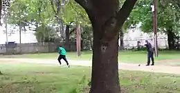 Video screenshot of Scott in a turquoise shirt and black pants running away to the left, many meters away, with Slager in uniform pointing his gun at Scott in the right of the screen