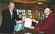 Prince Willem Alexander offers a waterbucket from the Royal Yacht "De Groene Draeck" Two senior officials watch.