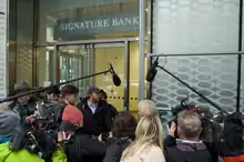 Reporters swarm a woman in white hair directly outside of an entrance to a bank.