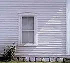 old white clapboard house, purplish hue case by shade, with window in center reflecting sky, stone base surrounded by weeds carefully rendered