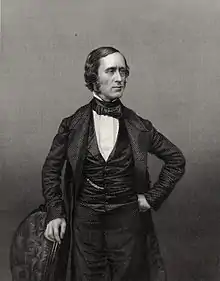 photograph of standing figure facing right, left hand on hip, right hand resting on chair