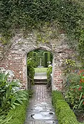 a view of an arch in a pink brick wall with a path leading to a statue