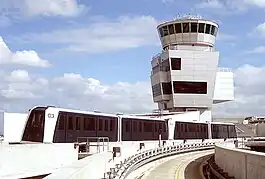 A four-car train traveling on top of a building with an empty concrete guideway in the foreground.  An airport control tower is in the background, in front of a blue, cloudy sky.