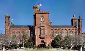 A brick building, reminiscent of a castle, slightly obscured by bare trees. There is a large central tower with the entrance at its base as well as smaller tower at each corner of the building, each with a varying design.