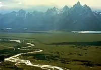 A braided light-colored stream winds across a flat grassy valley in the lower left while jagged gray peaks, laced with strips of snow, rise above a distant lake under a bright sky