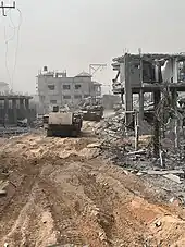 Two tanks roll on dirt, surrounded by a massive amount of destruction. There is rubble on the ground, and buildings that have been exploded