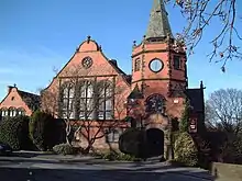 A brick building with stone dressings seen from the front.  At the centre is a bay with a large shaped gable containing a big window; to the right is a bay consisting of a turret containing a door and a clock face and a spire; to the left is a smaller bay with a shaped gable.