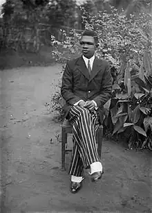 Black-and-white self-portrait of Alonge, dressed in formal clothes, sitting in a chair with his legs crossed at the knee