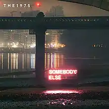 In a city at night under beside the bridge, a pink-colored neon sign reading "Somebody Else" with the band's name, "The 1975", in white text on the top left.