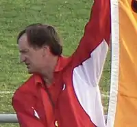 A dark-haired Caucasian man wearing a red and white jacket looks down and to his right whilst holding up a yellow flag, a small portion of which is visible in the frame.