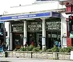 A entrance behind a raised bed of green plant and under a blue sign reading  "SOUTH KENSINGTON STATION" leads into an arcade of shops. Above the entrance there is a glass panel with white lettering reading "METROPOLITAN AND DISTRICT RAILWAYS" and "SOUTH KENSINGTON STATION".