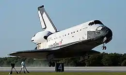 Columbia touches the concrete runway with its rear landing gear at Kennedy Space Center.  The tires leave smoke in their wake.  Green grass in front of the runway, trees behind, and the blue sky above complement the black and white orbiter.