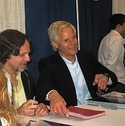 The picture shows several people sitting at a table. The one in the center has white hair and is looking up.