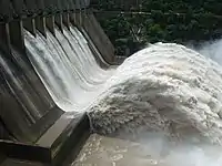 Aerial image of a river flowing from a concrete structure in the shape of a waterfall, with some forest in the background