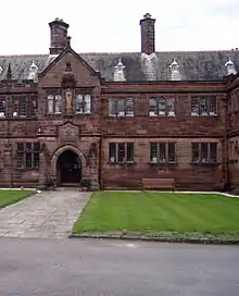 Part of a two-storey stone building seen directly from the front.  A paved path leads to a doorway in a slightly protruding bay; over the doorway is an inscribed plaque, and above that is a statue in a niche.