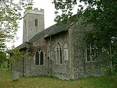 A small flint church seen between trees from the southeast, with a battlemented tower at the far end