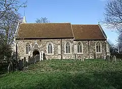 A plain stone church with a red tiled roof seen from the south, with the top of a small spire seen protruding above the left of the nave