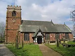 A red sandstone church seen from the south; on the left is a tower with a clock; the body of the church is to the right with a timber-framed porch. A path leads to the door in the porch with gravestones on each side