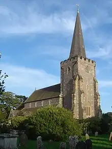 Three-quarter view of a stone church with a buttressed tower in the foreground. This has small battlements and a spire.  The nave roof, below which are four small, evenly spaced windows, is visible, but its aisle and an attached porch are obscured by a bush.  There are gravestones and a table tomb in the foreground.