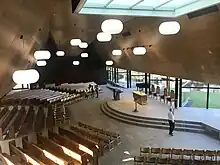 a low curved stage is backed by windows with pews around. white oval lamps float beneath the timber roof and interior panels