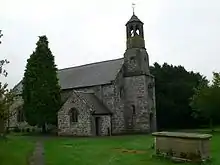 A small stone church seen from the northwest. On its right is a simple octagonal tower. Immediately in front of the church is a yew tree and in the foreground to the right is a chest tomb