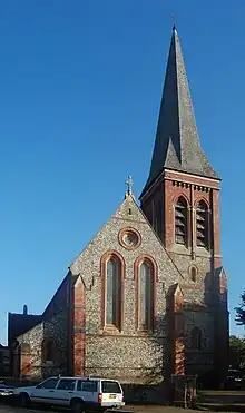 Side view of a long flint-built church with several parts, each with a different roofline.  A spire is visible behind the body of the church.  All windows are framed by brickwork.