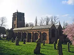 A substantial red sandstone church seen from the southeast. The tower and body of the church are crenellated. In the churchyard are gravestones and at the right extremity is a tree in flower