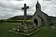 A plain greystone cross, in a mounting block, on top of four stone steps, with a small stone church in the background