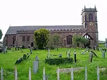 A red sandstone church seen from the north with a tower containing a clock face to the right. The nearest aisle is crenellated and in the churchyard in the foreground are gravestones, most of which are tilted