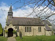 A small church seen from the south through the branches of a tree; directly ahead are the porch and a spirelet; to the right is the body of the church