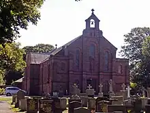 A cream, rendered brick building seen from the south, with dark slate roofs. The right third of the building (looking from the front) is on two storeys, and the remainder is on one.