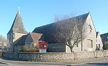 Three-quarter view of a long flint church with an extremely wide, stumpy tower on the left. This is topped with a dumpy grey spire.  The brown church roof is heavily discoloured.  A tree in full leaf obscures the near corner.