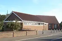 A long, low brick building in two non-aligned parts. Nearer the camera, in three-quarter view, is a hall with white soffits and window-frames; the windows are immediately below the roofline.  Behind it, in profile and mostly hidden, is a longer building of brown and red brick and with a steep, high roof.