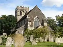 Rear view of a stone church with a castellated tower at the far end. The nearest side has very dark stone, two heavy buttresses and a three-light lancet window with trefoils.  Trees surround the church on all sides, and there are several gravestones in front.