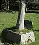 A very weathered square stone block with a short shaft leaning to the right