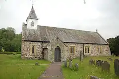 A small, simple, low church seen from the south. A small porch protrudes and to the left, and a small spire with a timber lower stage