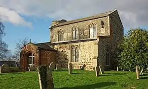 A photograph of a building with several windows standing on a field of green grass containing tombstones all under a blue sky with white clouds