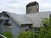 Very close view of a low iron church with a corrugated iron roof and a stone tower.  This has a low brown cap-style roof and a projecting crucifix.