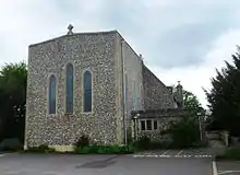 Front view of a flint church with a tall square façade topped with a small stone cross and with two round-headed windows flanking a taller, narrower one.  There is a paved parking area in front.