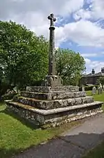 A stone cross on a chamfered column, on four stone square steps, with trees in the background