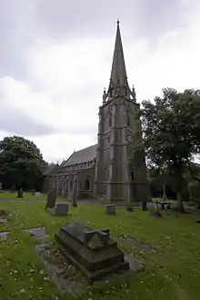 A church with a prominent spire seen from the northwest. Beyond the tower is the body of the church with a clerestory and north aisle. In the foreground are gravestones, the nearest one of which carries a sculpted cross